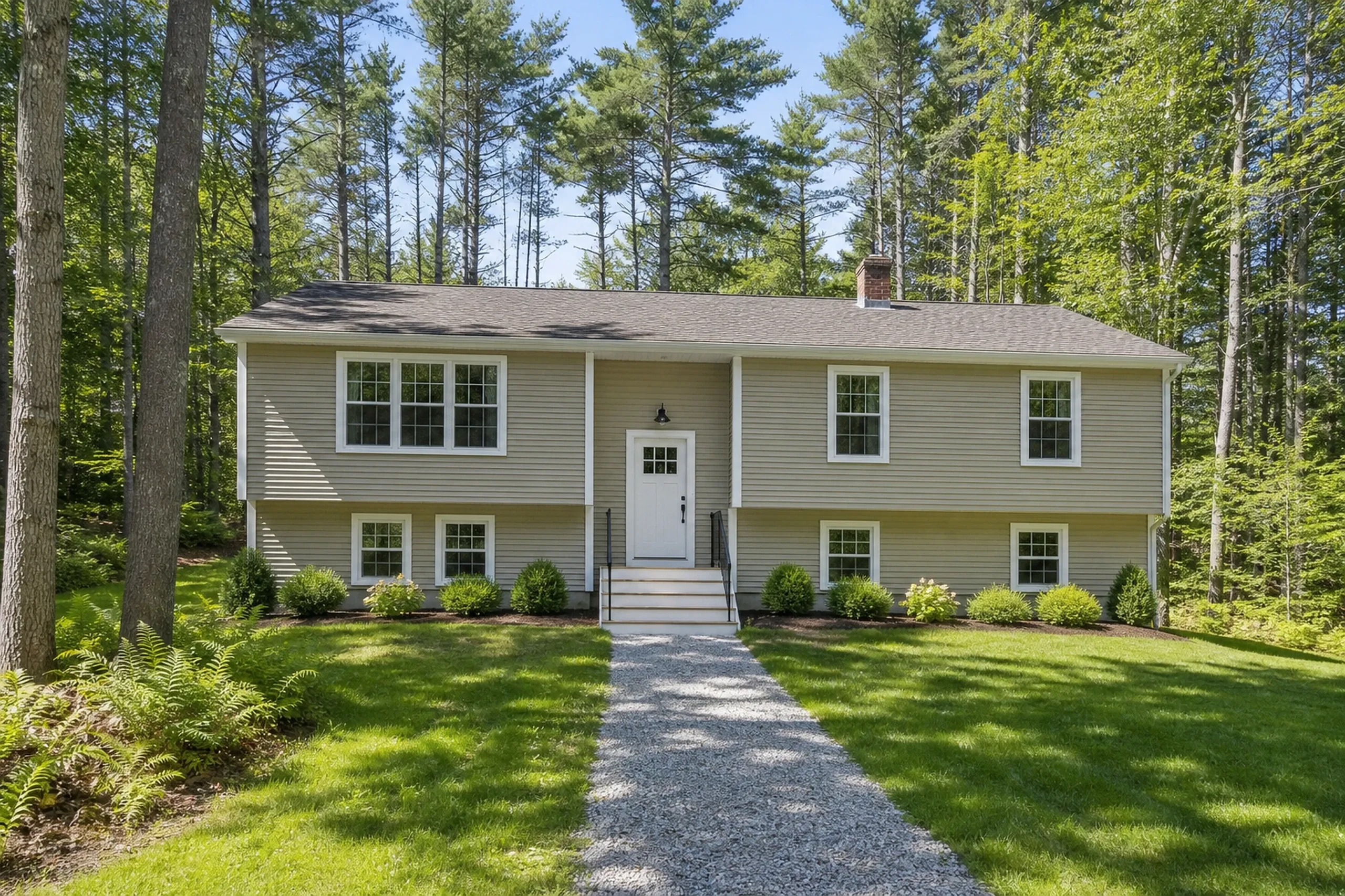 New vinyl siding in Southern Maine: a 1970s raised ranch on a wooded lot finished in tan Dutch-lap vinyl siding with white window trim and white corner posts, tall pines behind it and a gravel walk to the door, midsummer.