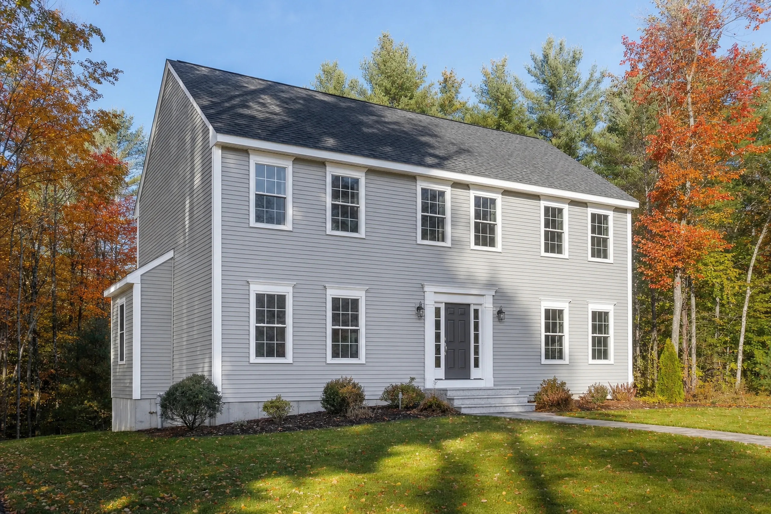 Siding installation in Gorham, ME: a two-story 1990s colonial on a wooded subdivision lot finished in new pale gray vinyl lap siding with crisp white trim, corner boards and window casings, early fall foliage