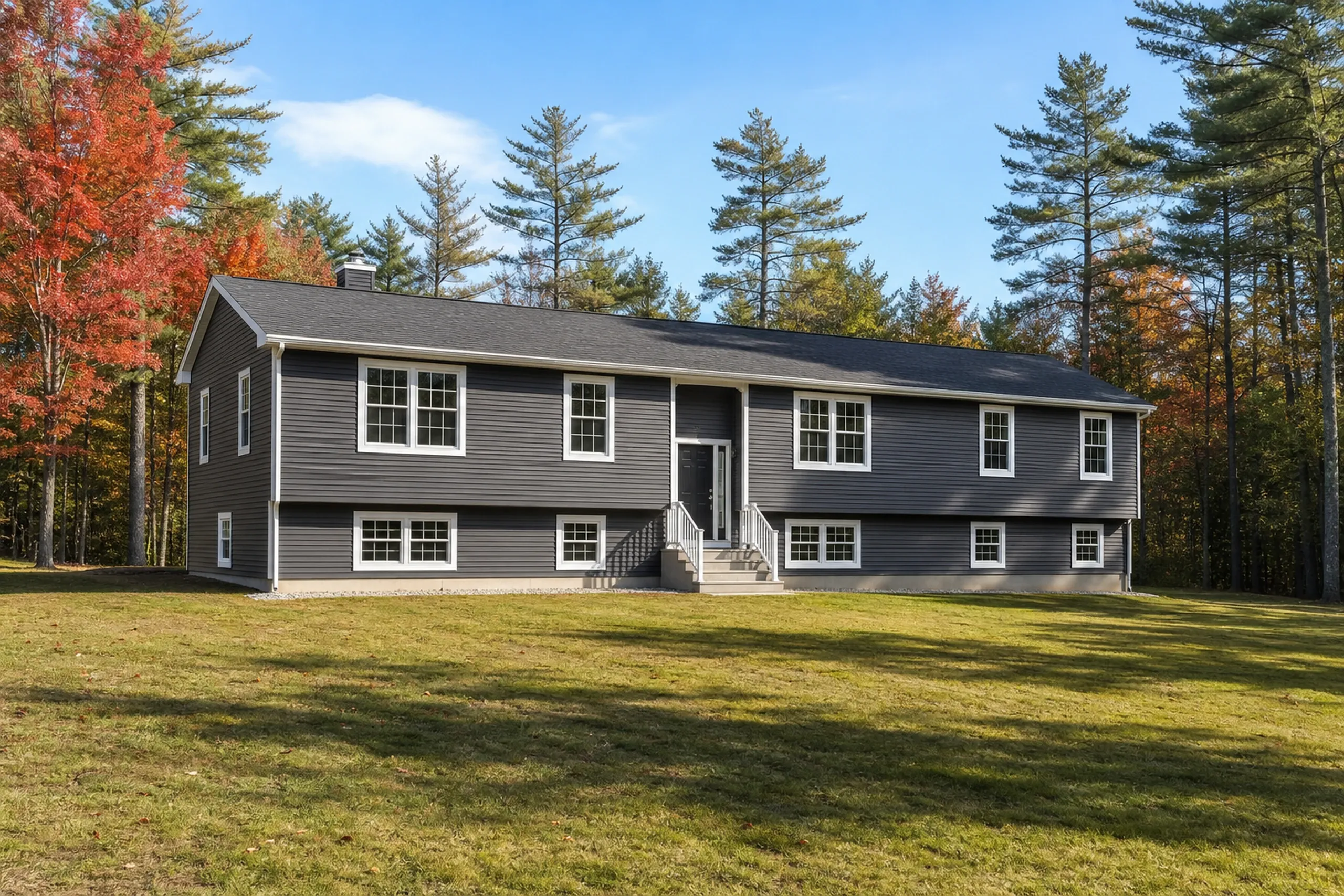 Windham re-sided raised ranch in early autumn: a re-sided raised ranch on a wooded Windham lot, deep slate-gray vinyl lap siding with white window trim and corner boards, a wide front lawn and red maples at the edge of the woods, crisp early-autumn light.