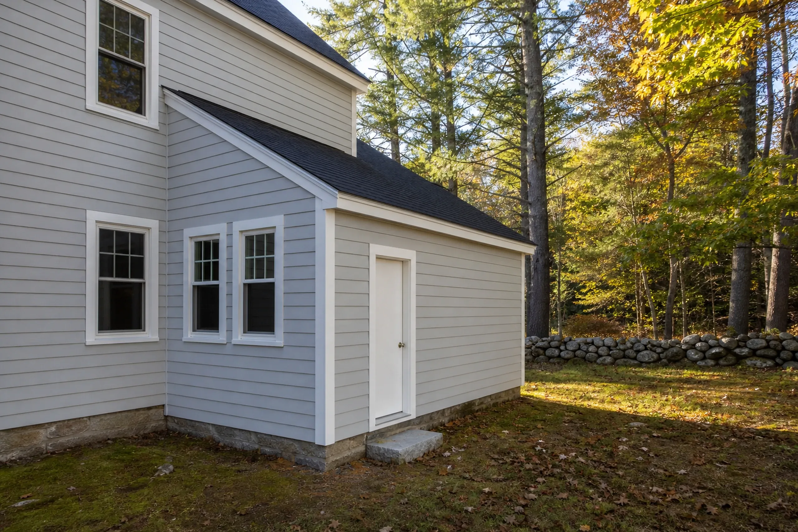 Yarmouth back wall: fiber cement lap siding painted pale gray on the rear ell of an older house, white corner board and window trim, a wooded back yard and no street in view, early fall.